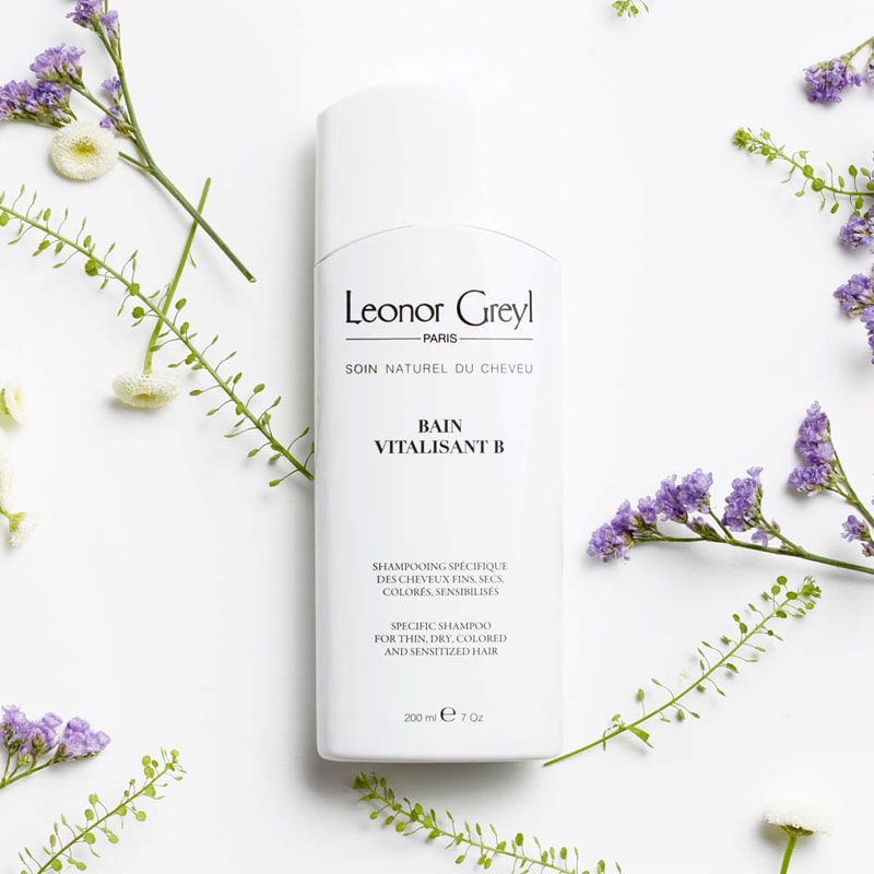 Lifestyle shot top view of Leonor Greyl Bain Vitalisant B Shampoo (200 ml) with purple and white flowers in the background