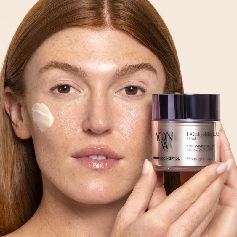 Model holding Yon-Ka Paris Excellence Code Creme (50 ml) with product smear on cheek to show color and texture