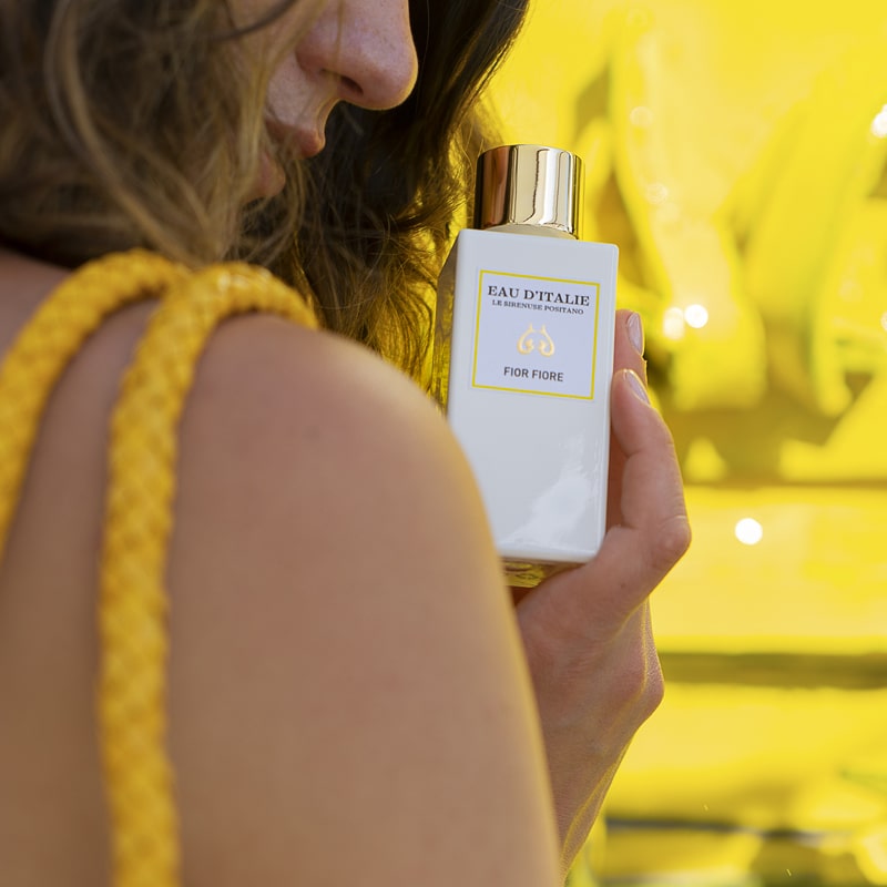 Close up shot of model holding a bottle of Eau d'Italie Fior Fiore Eau de Parfum Spray (100 ml) with bright yellow ornate sculpture in the background