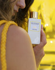 Close up shot of model holding a bottle of Eau d'Italie Fior Fiore Eau de Parfum Spray (100 ml) with bright yellow ornate sculpture in the background