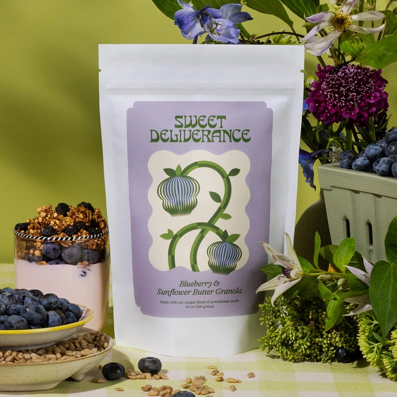 Lifestyle shot of Sweet Deliverance Blueberry &amp; Sunflower Butter Granola (9.5 oz) bag with granola in yogurt and purple flowers and green leaves in the background
