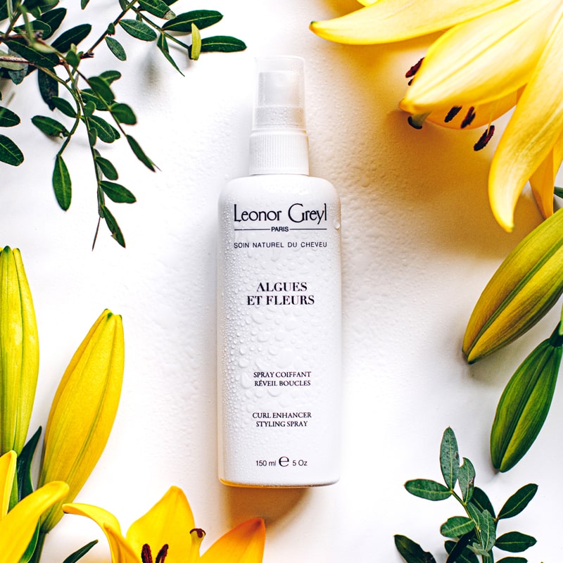 Lifestyle shot of Leonor Greyl Algues et Fleurs Conditioner Curl Enhancer Styling Spray (150 ml) with yellow flowers and leaves in the background