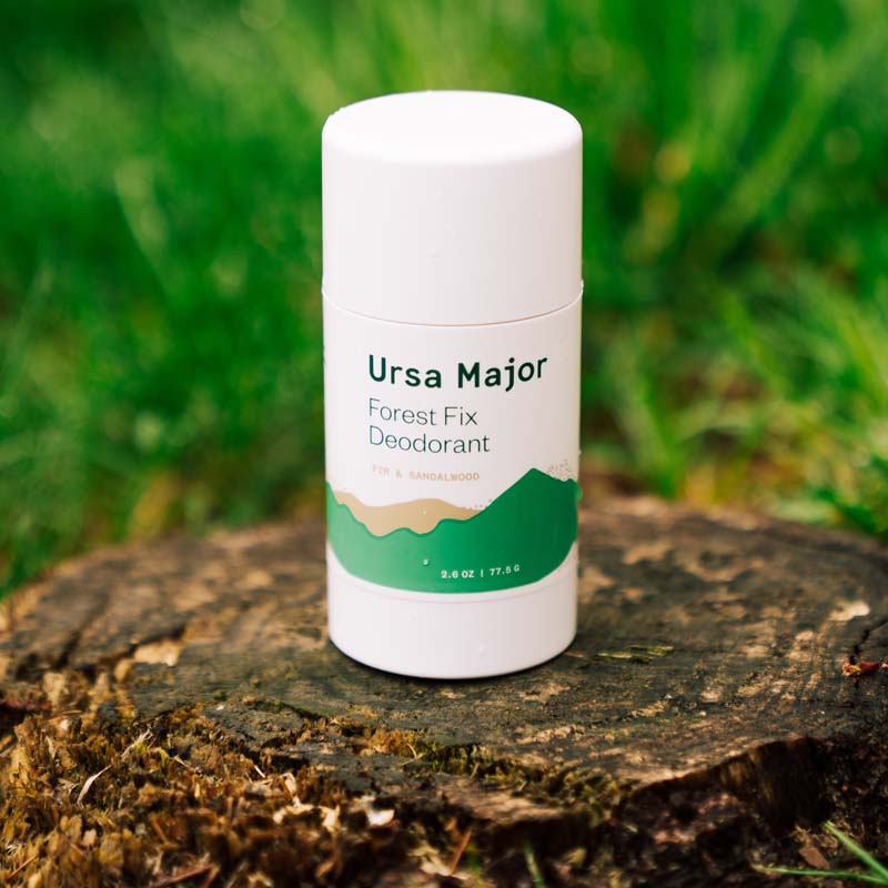 Lifestyle shot of Ursa Major Forest Fix Deodorant (2.6 oz) on tree stump and grass in the background