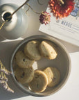 Lifestyle shot of Makabi & Sons Rose Pistachio Cardamom Cookies - Maroc, 6 cookies shown in a dish with box and vase with flowers in the background