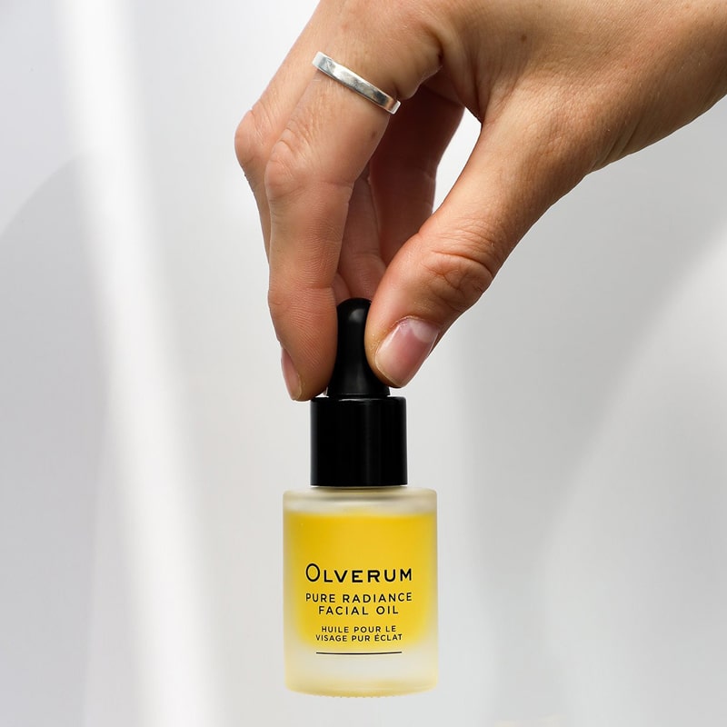 Olverum Pure Radiance Facial Oil with model&#39;s hand grasping top of dropper on closed bottle