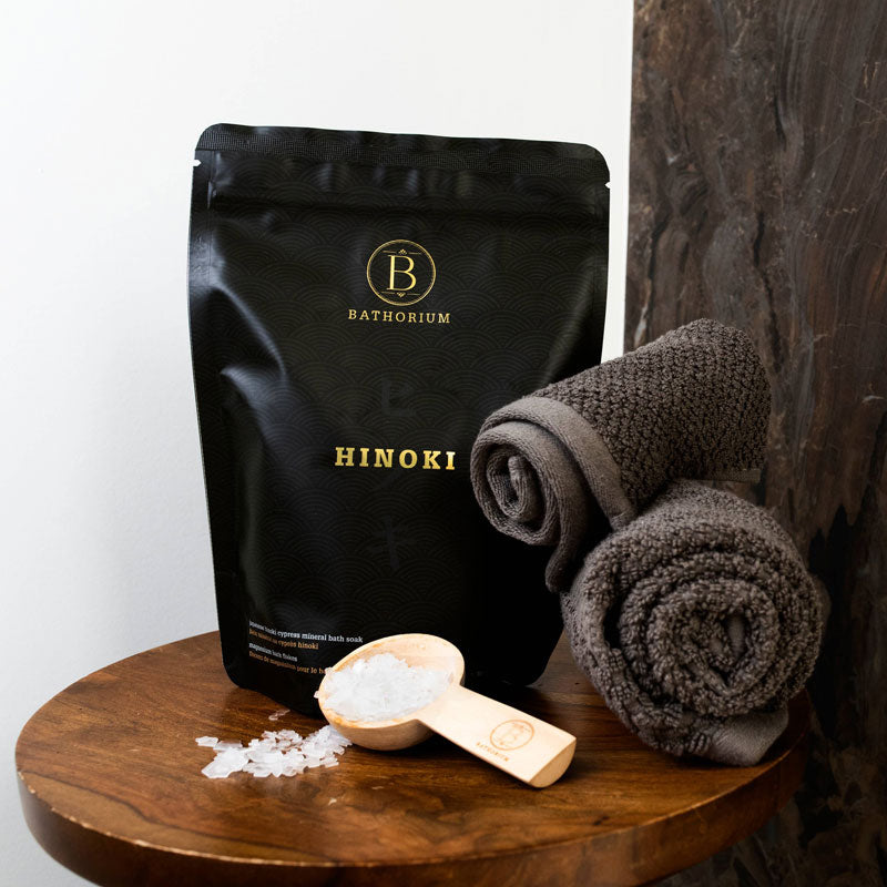 Bathorium Hinoki Magnesium Bath Flake Soak lifestyle setting with towel rolls and a scoop of bath flakes