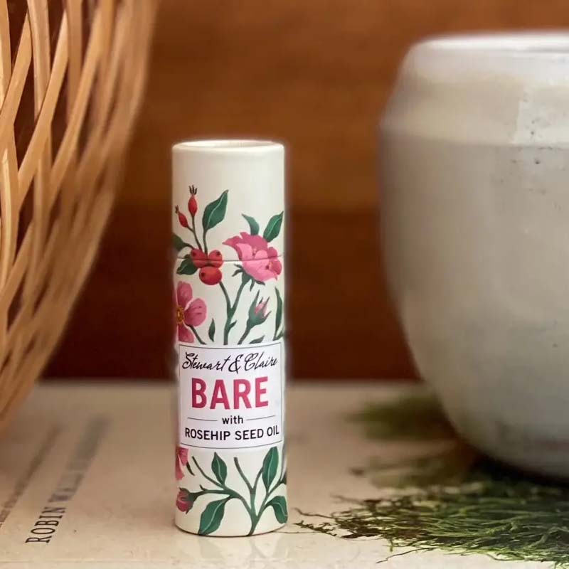 Stewart & Claire Bare Unscented Lip Balm with Rosehip Seed Oil showing on a book with a basket next to it