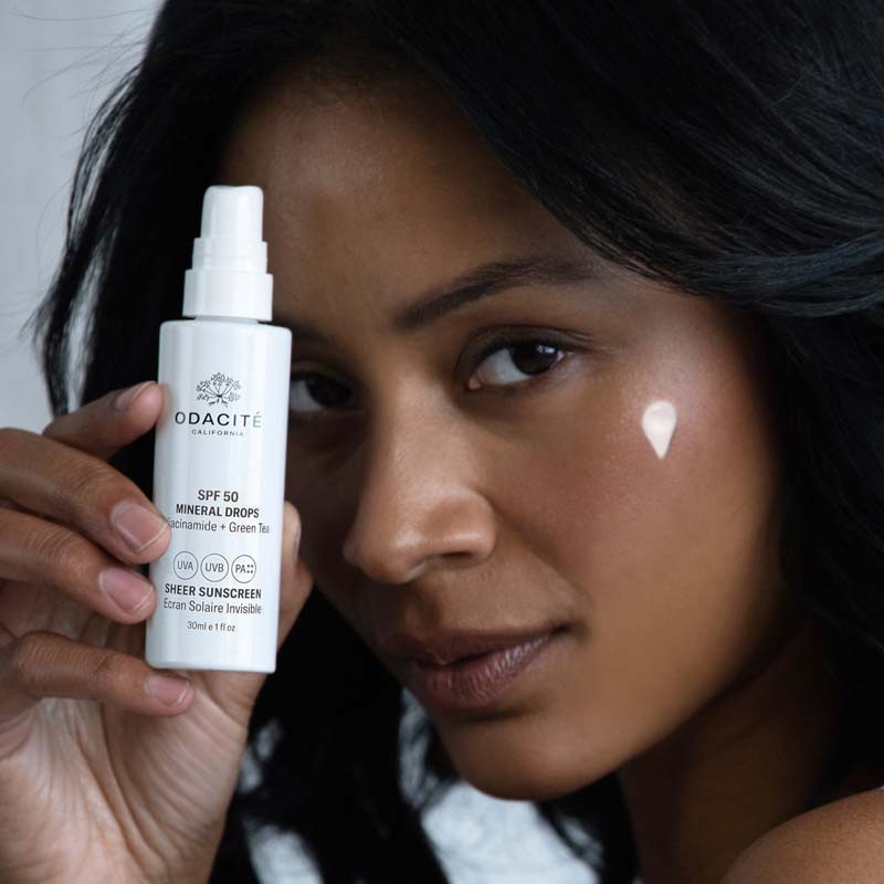 Close up of model holding Odacite SPF 50 Mineral Drops Sheer Sunscreen (30 ml) with smear applied to cheek