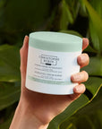 Christophe Robin Hydrating Cream Scrub shown in models hand with green leaves behind