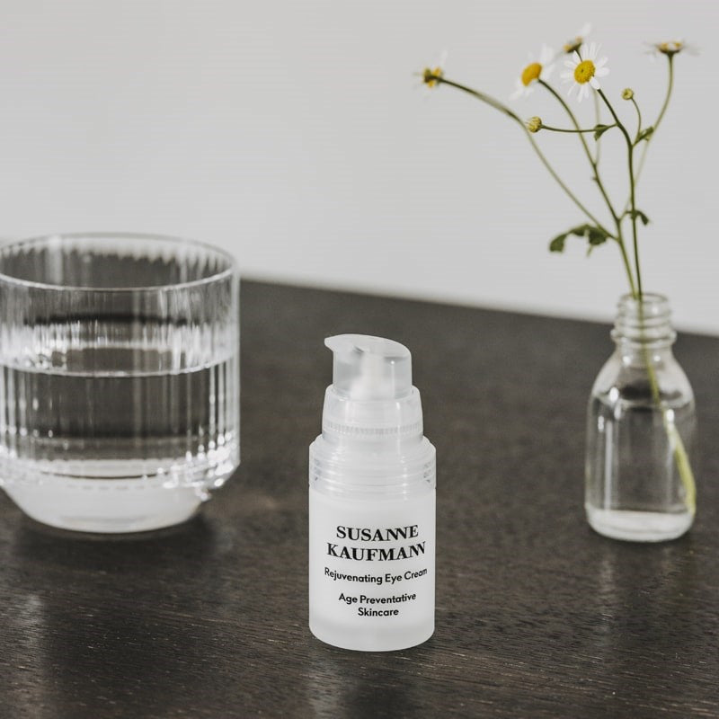 Susanne Kaufmann Rejuvenating Eye Cream lifestyle shot of product on wood table with a glass of water and a small vase with flowers in it