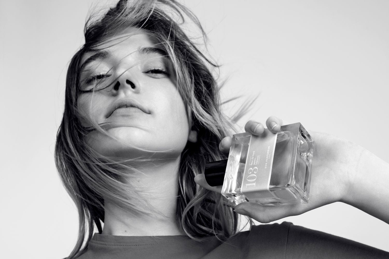 A black and White image of young woman with hair blowing in the wind holding a bottle of Bon Parfumeur Paris 103 Tiare Flower Hibiscus Jasmin