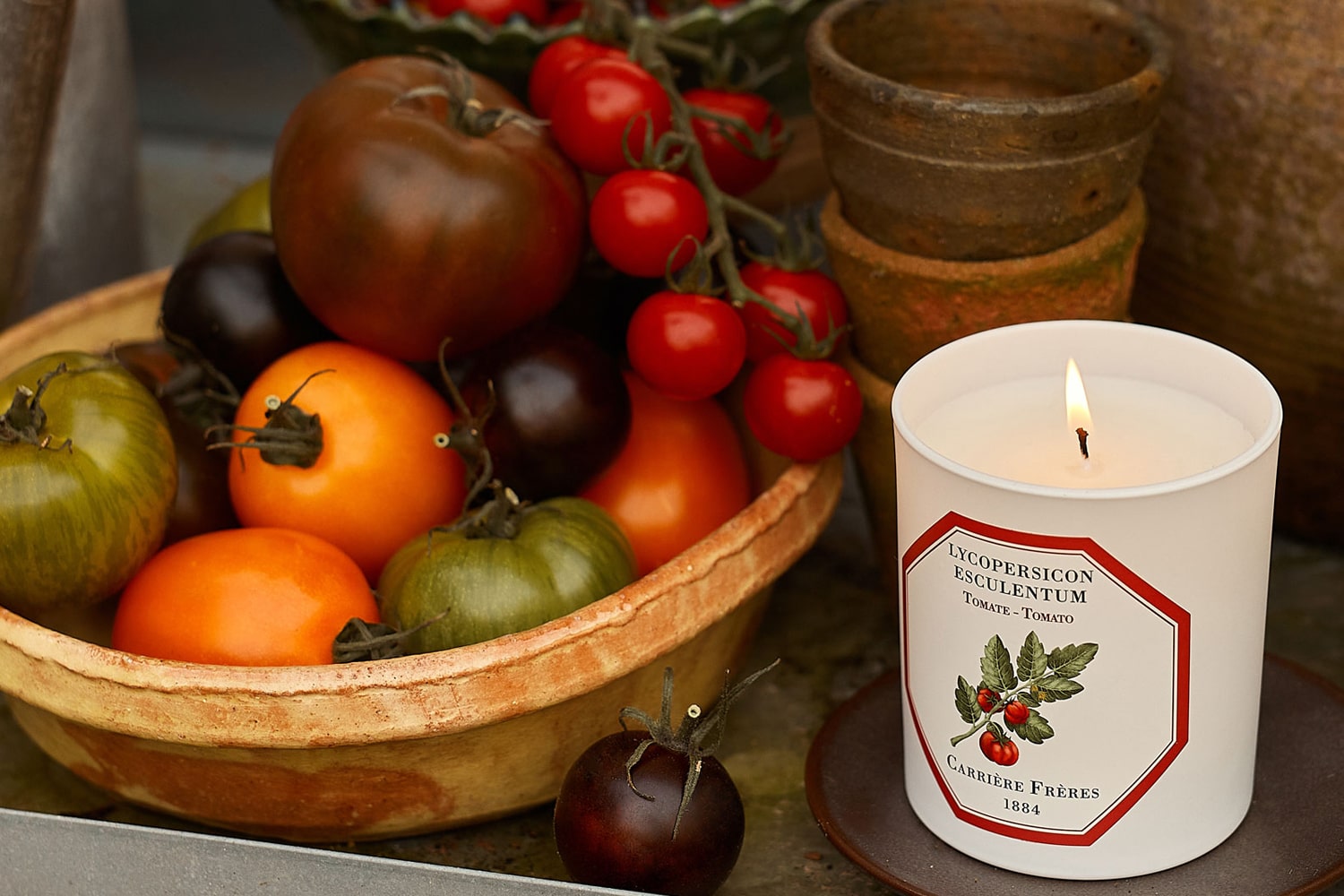 Lifestyle shot of Carriere Freres Tomato Candle with a basket of a variety of tomatoes next to candle