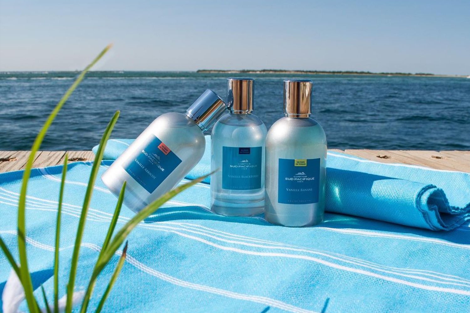 Lifestyle shot of A selection of Comptoir Sud Pacifique fragrance bottles on blue and white stripe towel with beach in the background