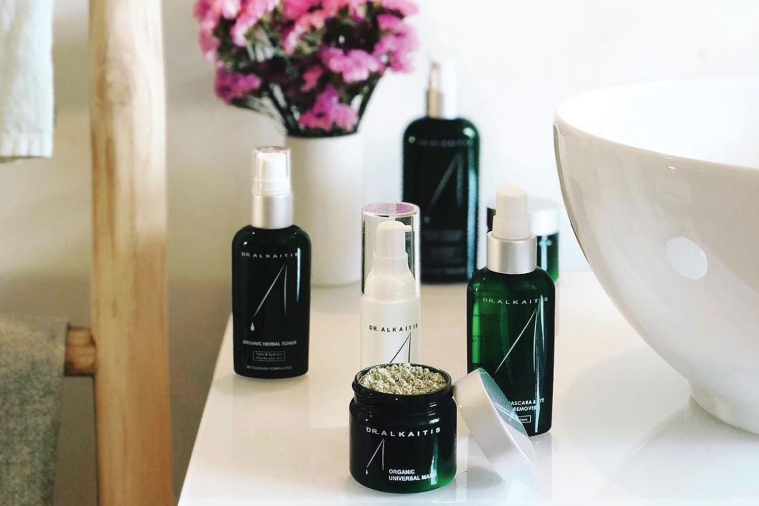 A selection of Dr. Alkaitis skincare products next to a raised bowl sink and vase with pink flowers in the background