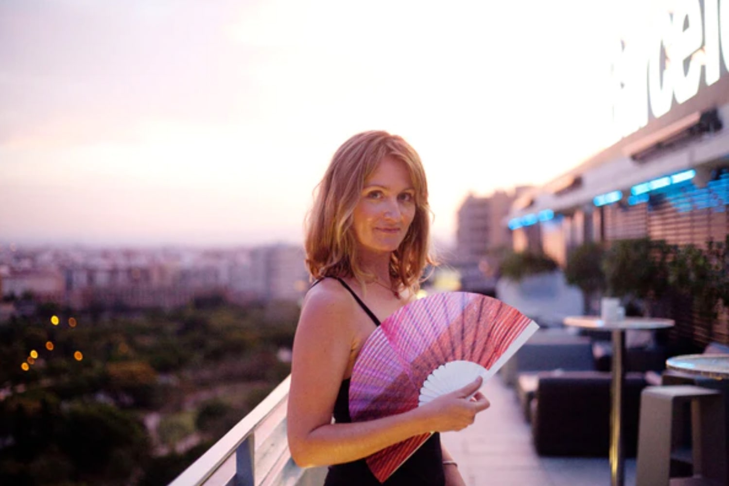 Founder of Khu Khu, Victoria Speyer holding fan on an outside balcony overlooking a cityscape