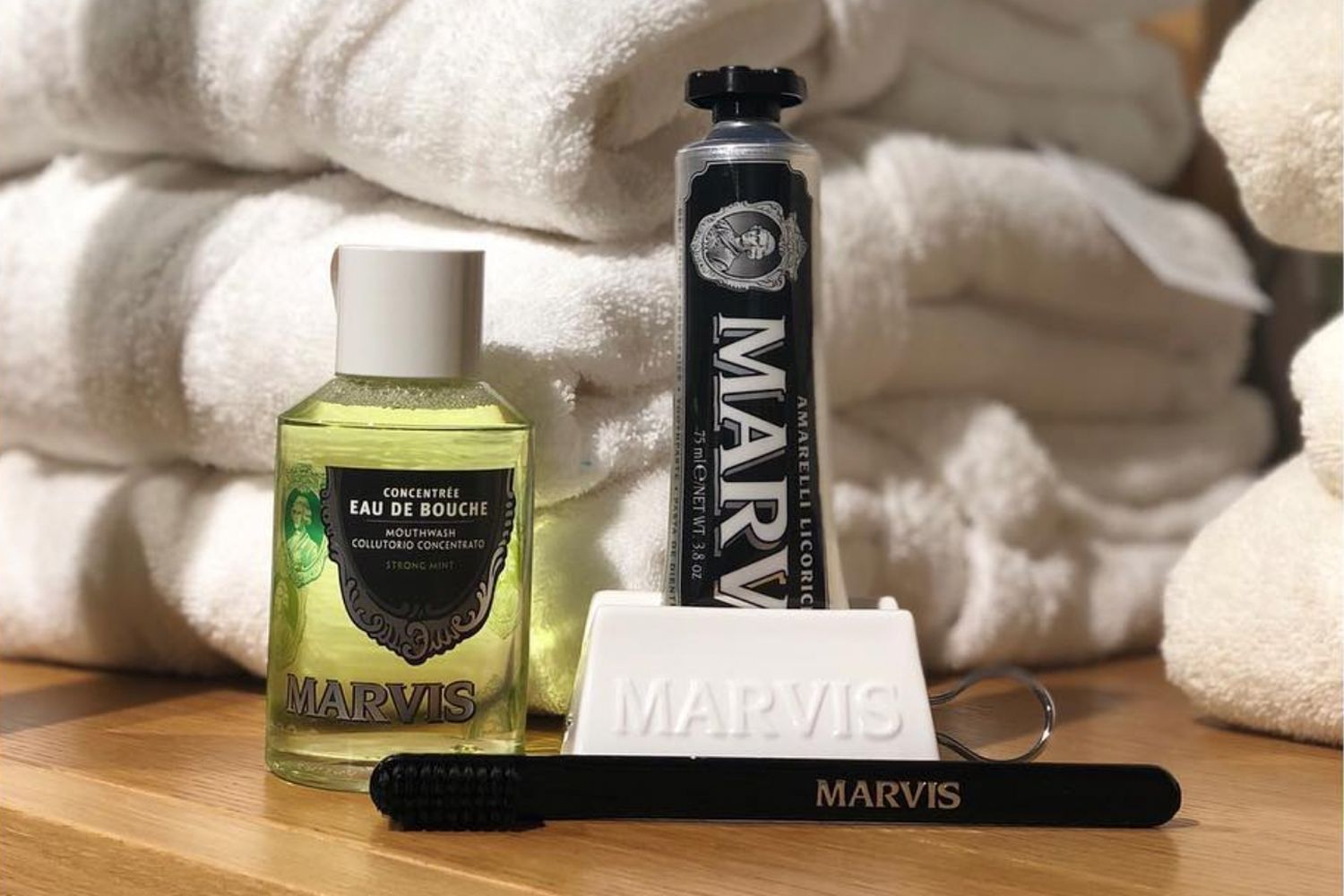Beauty shot of Marvis mouthwash, toothpaste and ceramic toothpaste dispenser with pile of folded white bath towels in the background