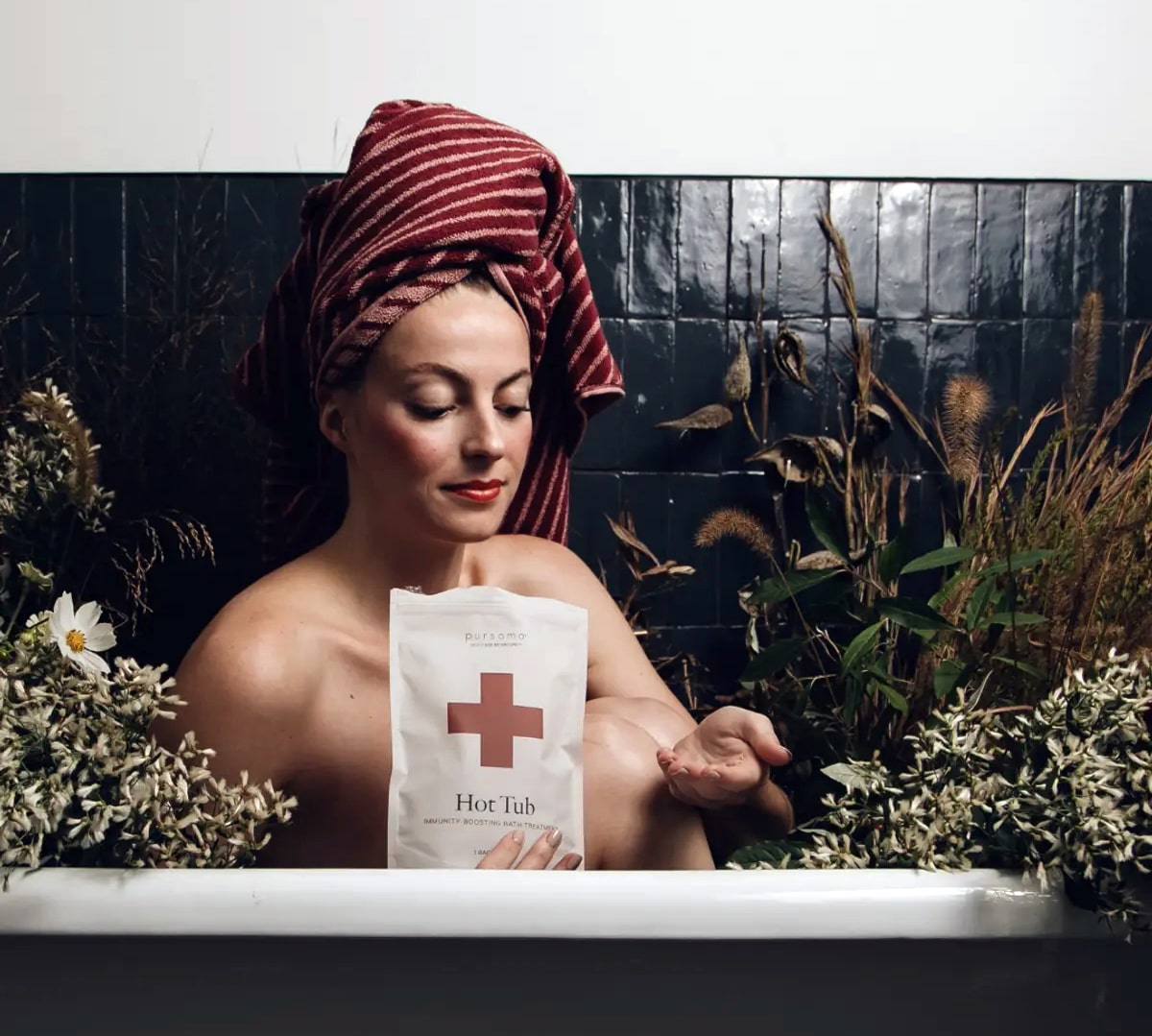 Lifestyle shot of model in tub holding a bag of Pursoma Hot Tub Bath Treatment surrounded by flowers