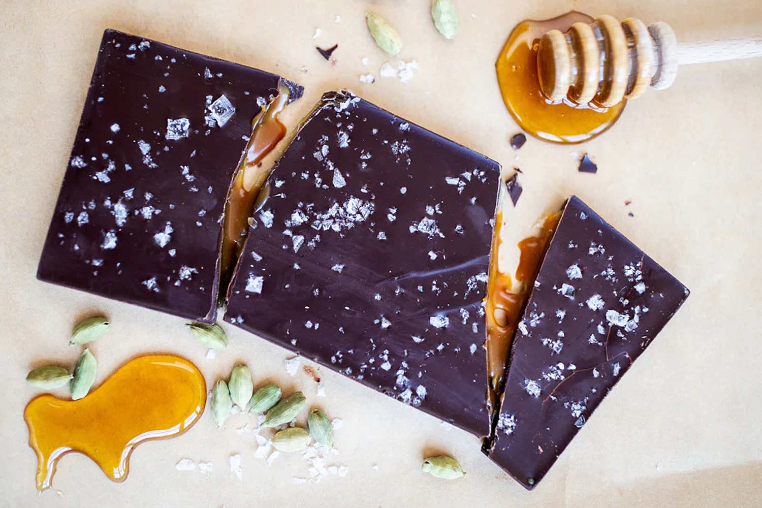 Lifestyle shot of Wildwood Chocolate Cardamom and Honey Caramel with Sea Salt chocolate bar with honey, cardamom and sea salt in the background and bar broken in pieces to show caramel inside