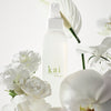 Kai Fragrance Body Glow Spray (4 oz) surrounded by white flowers on a light background