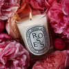 Diptyque Roses Candle - candle shown burning with pink roses in the background