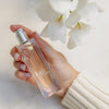 Hand holding Kai Eau de Parfum Spray (1.7 oz/ 1 oz) with white flowers in the background
