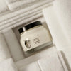 Lifestyle shot of Diptyque Rich Butter For The Body (200 ml) with folded white towels in the background