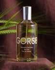 Lifestyle shot of Laboratory Perfumes Gorse Eau de Toilette on dark brown linen cloth with green stalks in the background