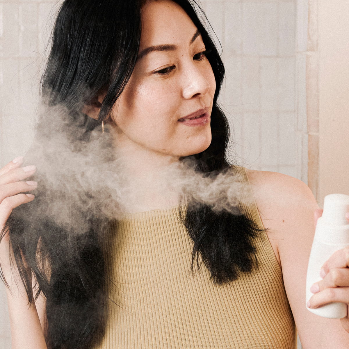 A woman with long black hair sprays Rahua Voluminous Dry Shampoo (51 g) onto her hair, creating a visible powder mist