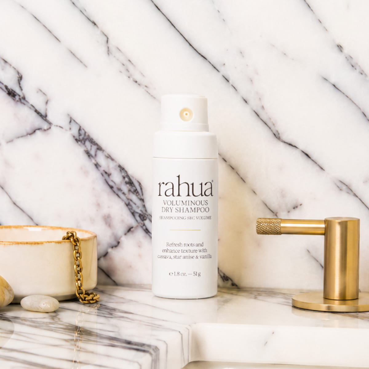 A bottle of Rahua Voluminous Dry Shampoo (51 g) standing on a white marble surface with a matching marble wall background, styled with a gold faucet, a small bowl, and stones