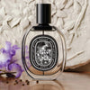 Fleur de Peau Eau de Parfum (75 ml) - product shown against stone wall with purple flower. 