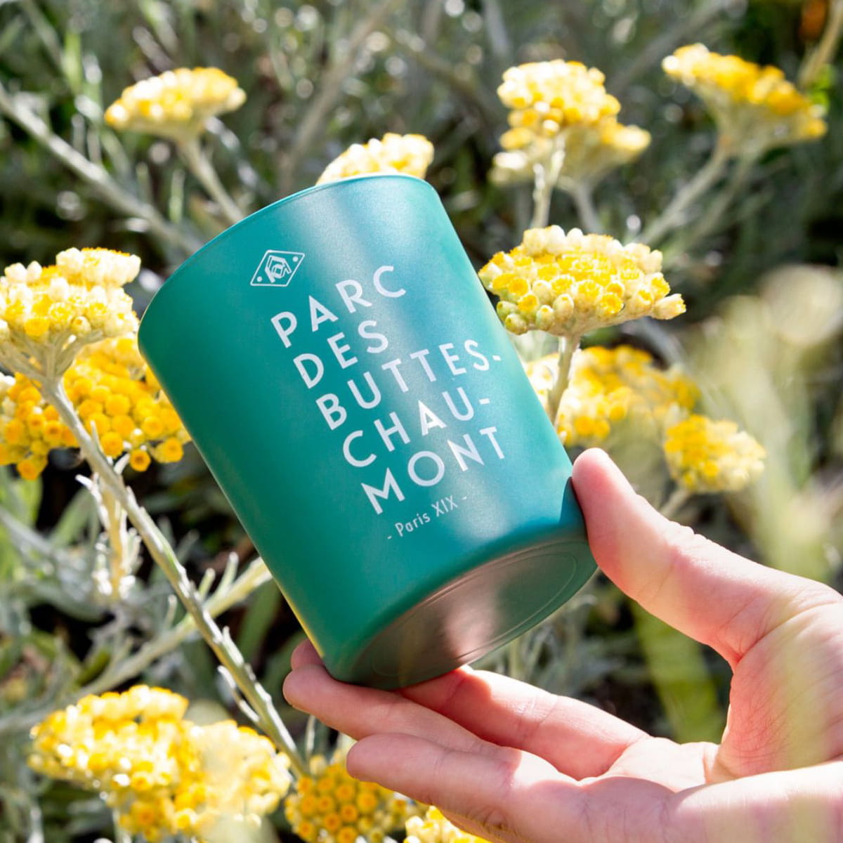 Kerzon Fragranced Candle - Parc des Buttes-Chaumont (190 g) - product shown being held in front of flowers.