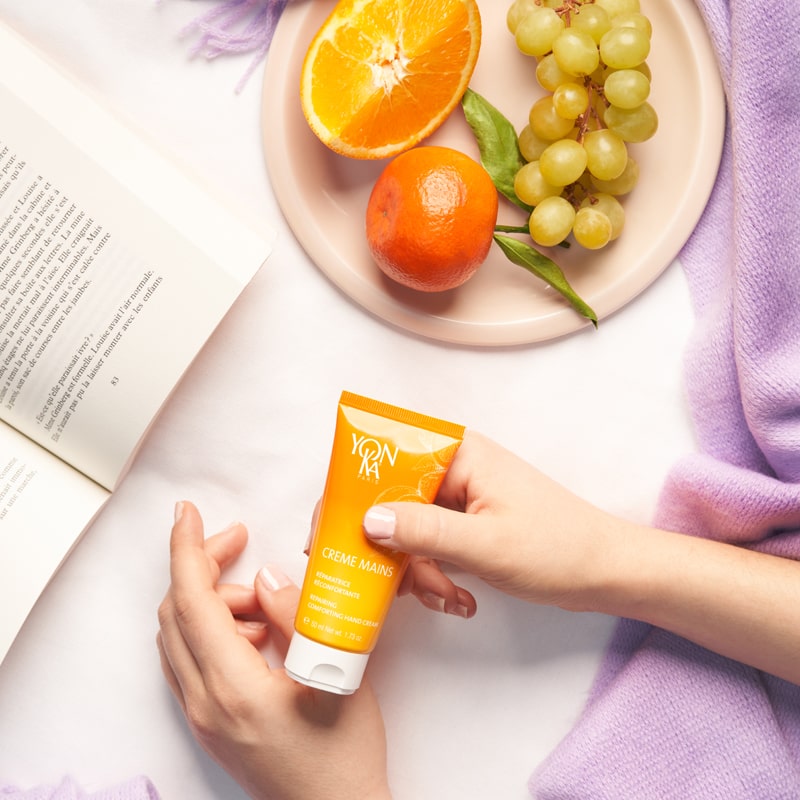Lifestyle shot top view of model holding Yon-Ka Paris Creme Mains - Vitalite Sweet Orange (50 ml) with opened book and plate of fruit in the background