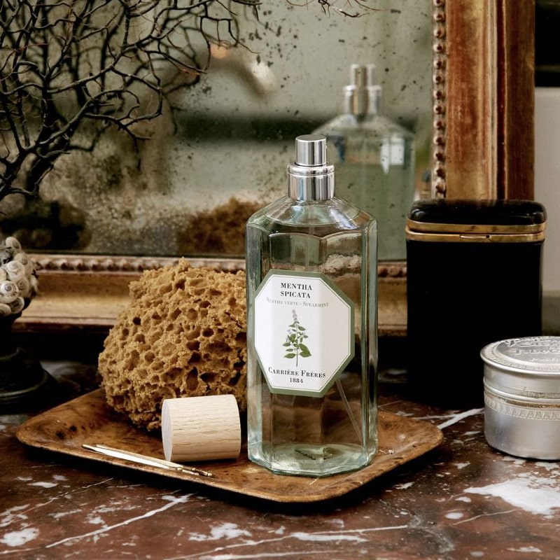 Lifestyle shot of Carriere Freres Spearmint Room Spray (200 ml) with cap off on small wood tray on marble table and mirror in the background