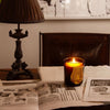 Trudon Cire Candle (9.5 oz) burning on an open book with a lamp and newspaper in the background