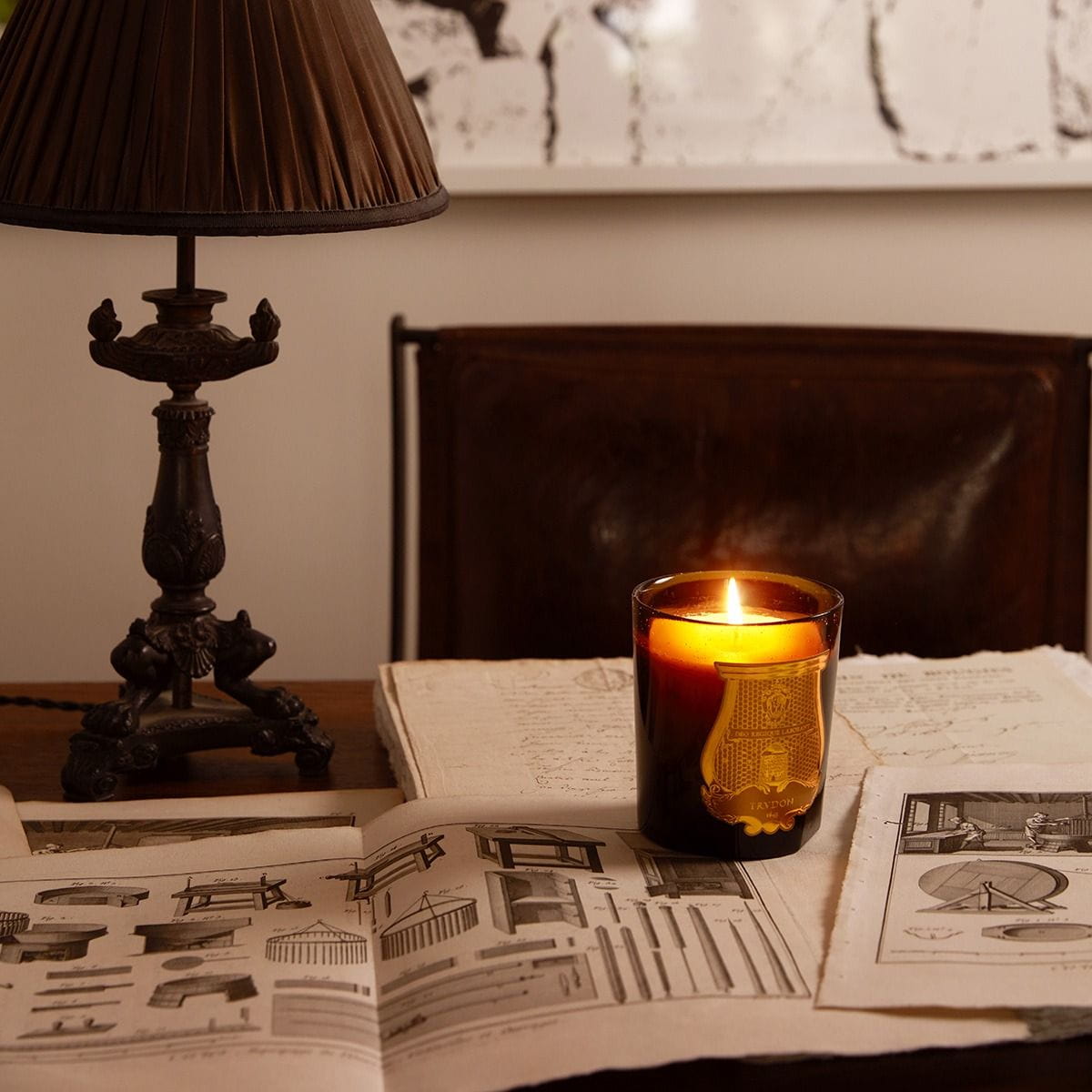 Trudon Cire Candle (9.5 oz) burning on an open book with a lamp and newspaper in the background
