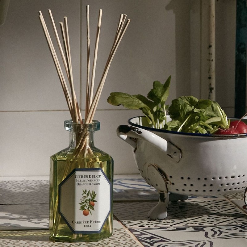 Lifestyle shot of Carriere Freres Orange Blossom Diffuser with reeds on decorative tile with strainer and vegetables in the background