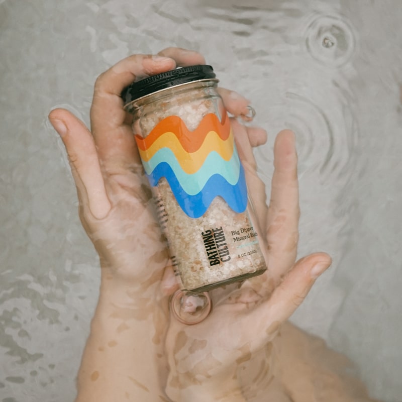 Model holding Bathing Culture Big Dipper Mineral Bath (8 oz) jar in hands in water