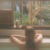 Model sitting in bath looking our window with tree and waterfall in the background and Bathing Culture Big Dipper Mineral Bath (8 oz) on window sill