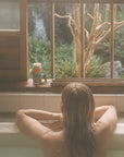 Model sitting in bath looking our window with tree and waterfall in the background and Bathing Culture Big Dipper Mineral Bath (8 oz) on window sill