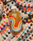 Close up shot of back side of Bathing Culture Refillable Rainbow Glass Mind and Body Wash in Cathedral Grove (16 oz) bottle with Bathing Culture Cosmic Rainbow (deadstock) body towel in the background