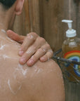 Close up of model applying Bathing Culture Refillable Rainbow Glass Mind and Body Wash in Cathedral Grove to shoulder with lather