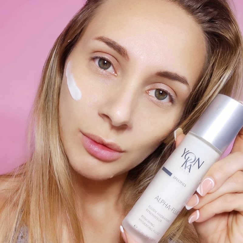 Model holding bottle of Yon-Ka Paris Alpha Fluid (50 ml) and product smear on cheek to show color and texture