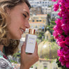 Lifestyle shot of model holding Eau d'Italie Eau de Parfum Spray (100 ml) with bougainvillea and Positano landscape in the background
