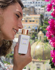 Lifestyle shot of model holding Eau d'Italie Eau de Parfum Spray (100 ml) with bougainvillea and Positano landscape in the background