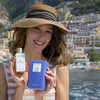 Lifestyle shot of model holding Eau d'Italie Un Bateau Pour Capri Eau de Parfum Spray (100 ml) and box with the Hotel Le Sirenuse and Positano cliffside in the background 