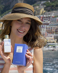 Lifestyle shot of model holding Eau d'Italie Un Bateau Pour Capri Eau de Parfum Spray (100 ml) and box with the Hotel Le Sirenuse and Positano cliffside in the background 