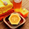 Lifestyle shot of Honey House Naturals Large Bee Bar Lotion - Natural (2 oz) on wood tray with honey in a glass jar and candle burning with folded towels in the background