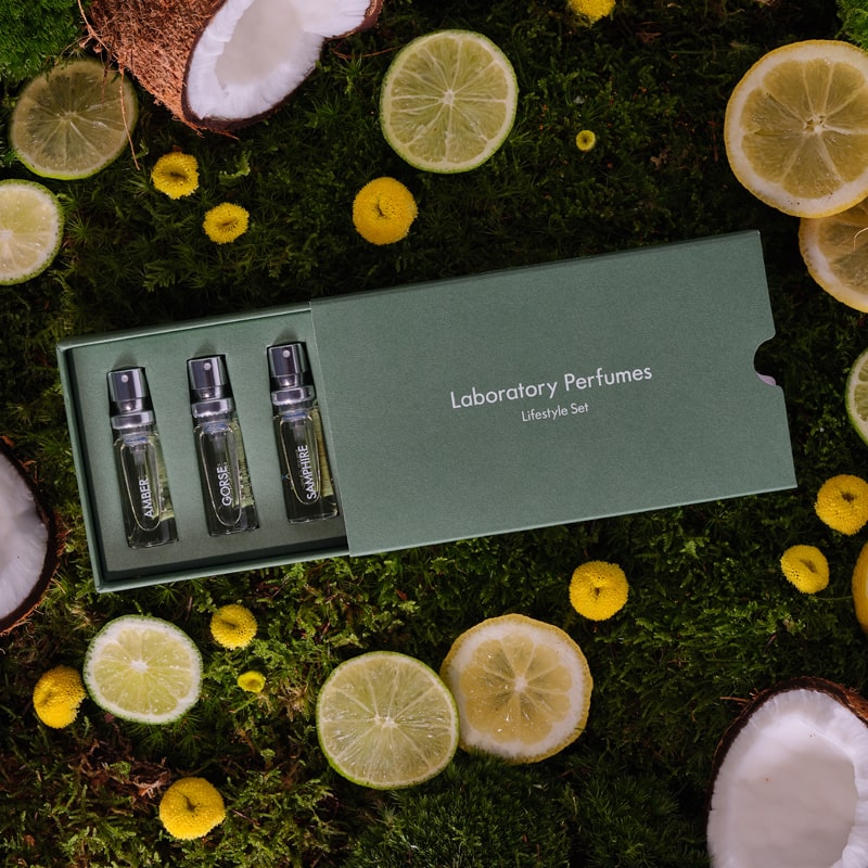 Lifestyle shot top view of Laboratory Perfumes Lifestyle Set on grass with box slightly open and yellow flowers, coconut, lemon and lime slices in the background