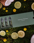 Lifestyle shot top view of Laboratory Perfumes Lifestyle Set on grass with box slightly open and yellow flowers, coconut, lemon and lime slices in the background