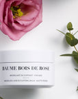 Lifestyle shot top view of Leonor Greyl Baume Bois de Rose with pink flower and green leaves in background